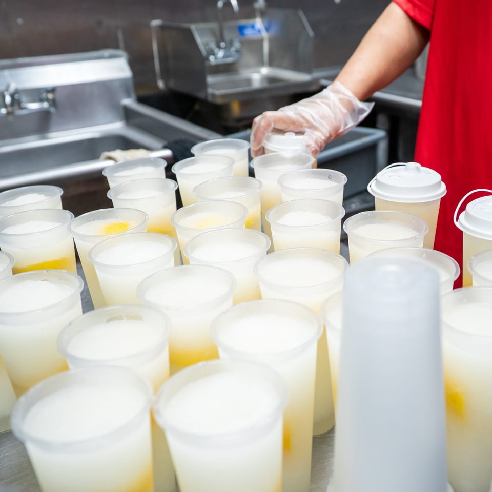 A gloved hand places a lid on one of many plastic cups filled with a light-colored beverage.