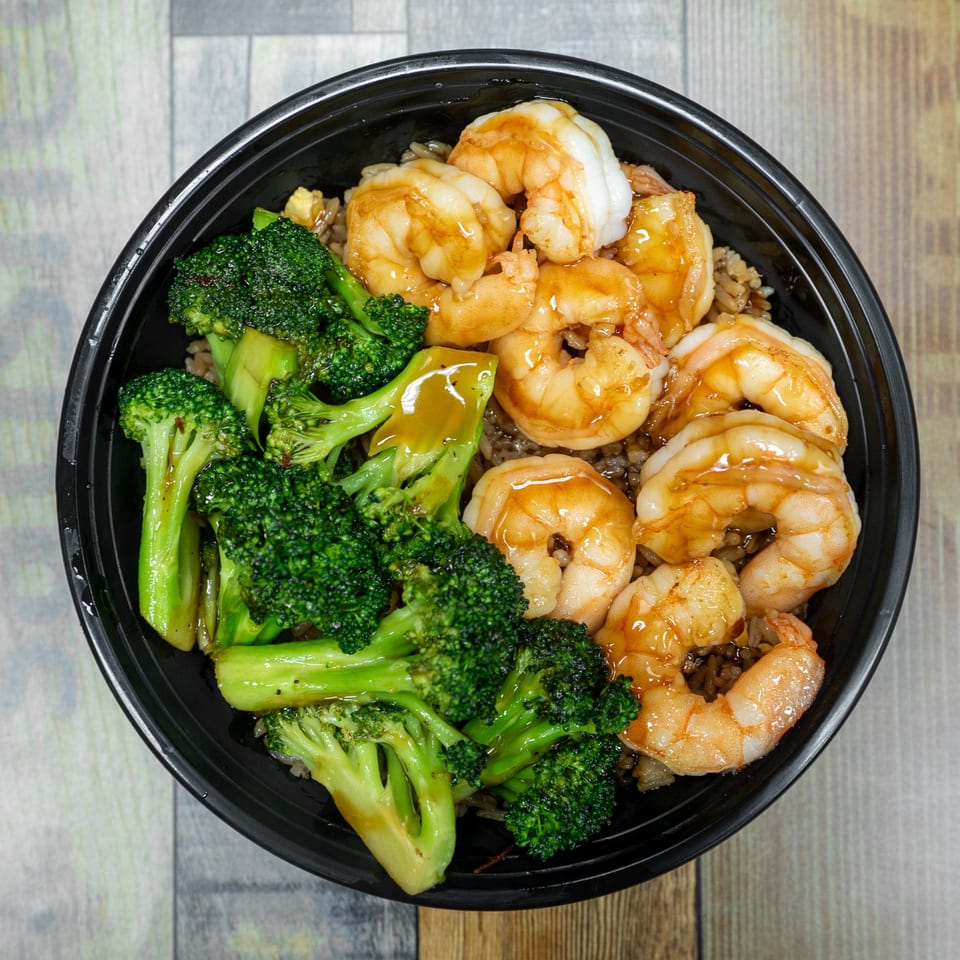 Top-down view of a black bowl filled with shrimp, broccoli, and rice, drizzled with sauce.