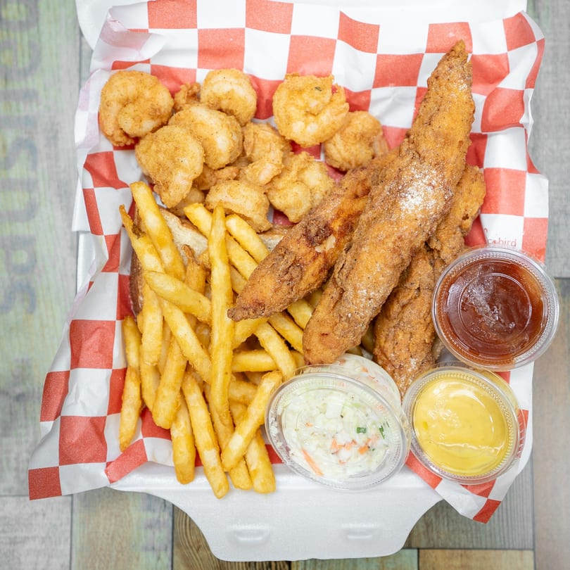 Fried fish and shrimp combo with fries, coleslaw, and dipping sauces in a takeout container.