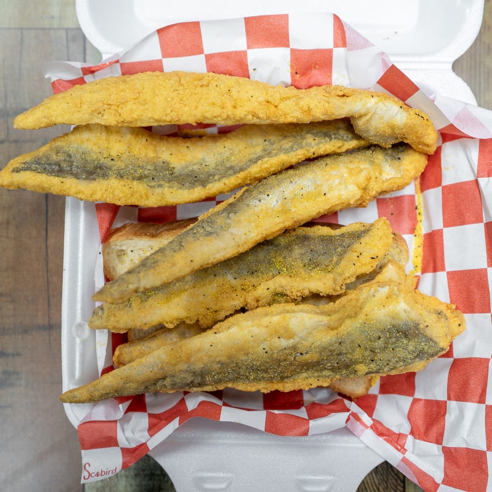 Crispy fried fish fillets with seasoning, served in a white styrofoam container with red and white checkered paper.