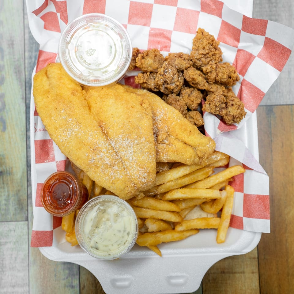Fried fish fillets, popcorn shrimp, and french fries with dipping sauces in a takeout container.